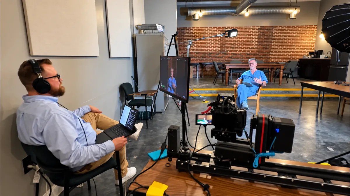 Trevor Chapman, wearing headphones with a laptop on his lap, monitors an interview in a brick-walled room. Across the room a man in blue scrubs speaks from a chair, and a monitor beside the camera slider shows him on screen.