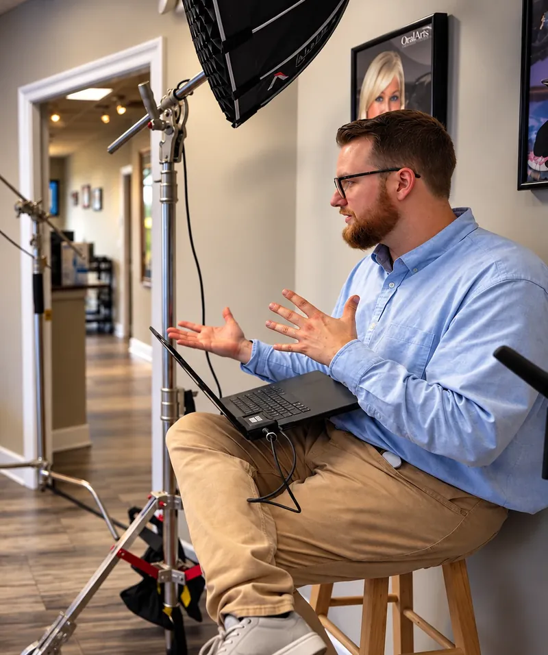 Trevor Chapman, bearded and in glasses and a light-blue shirt, sits on a stool with a laptop on his lap and gestures with both hands as he explains something on set, under a softbox light.