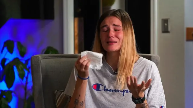 A woman in a grey T-shirt, seated for an interview, holds a tissue and is visibly emotional.