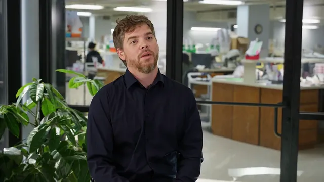 A man in a dark shirt is interviewed in a dental lab.