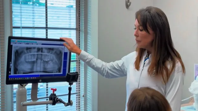A dentist in a white coat points to a panoramic dental X-ray on a monitor while a patient watches.