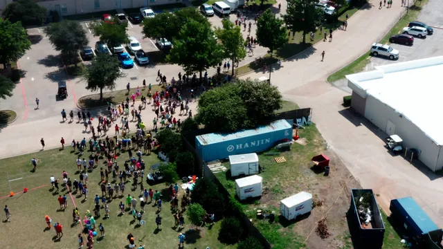 Aerial view of a crowd on a lawn beside a building and a parking lot.