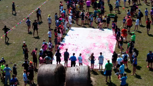 Aerial view of a crowd gathered around a large painted tarp on a grass field.