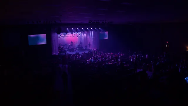 A wide view from the back of a dark hall toward a purple-lit stage and band.