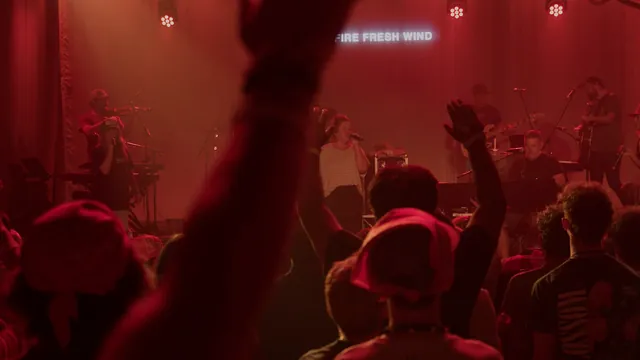 A band performs on a red-lit stage while the crowd raises their hands.