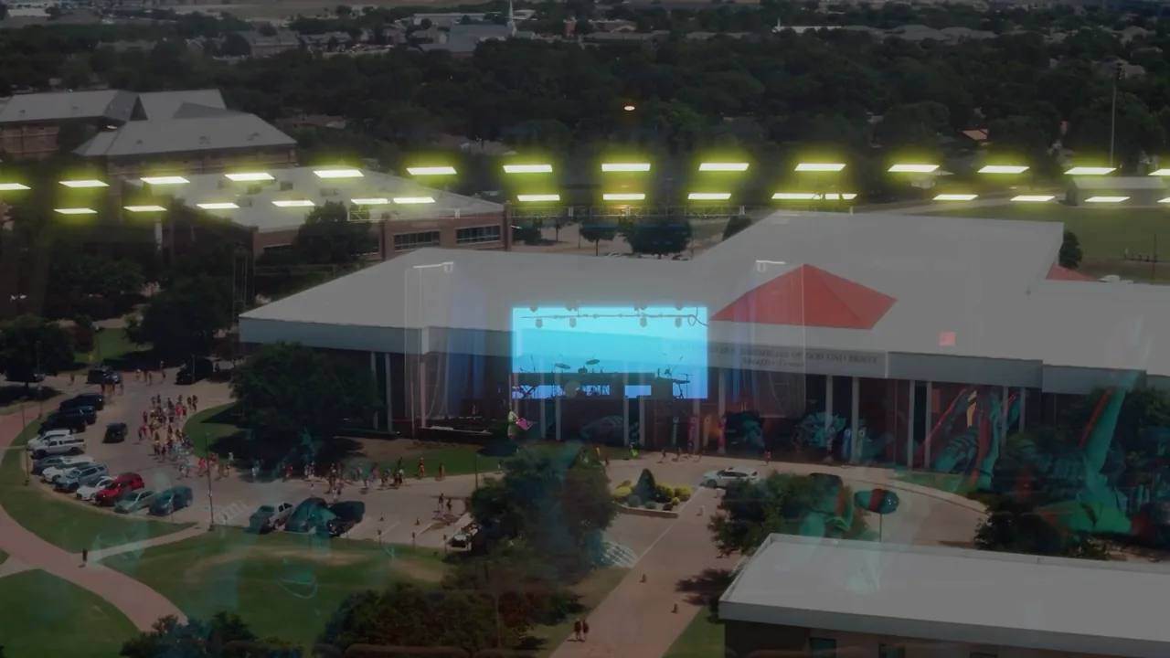 Aerial view of a campus event venue: a long white-roofed building with a lit screen at its entrance, with trees around it.