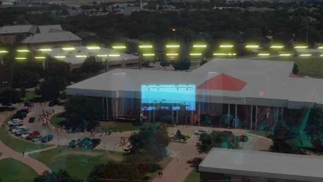 Aerial view of a campus event venue, a long white-roofed building with a lit screen at its entrance and trees around it.