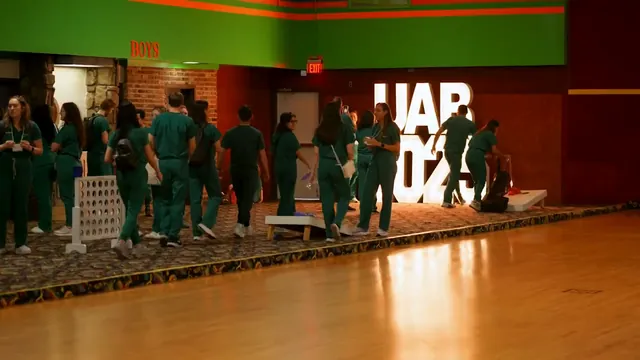 Students in green scrubs walk past large illuminated letters in an event hall with a wooden floor.