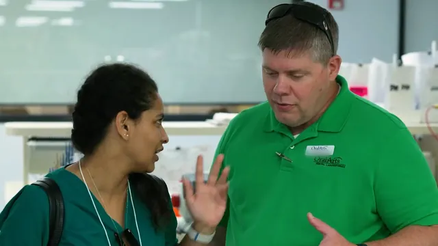 A lab staff member in a green polo explains something to a student.